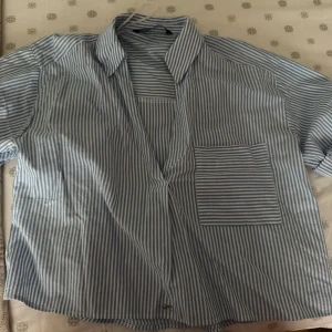 Blå- och vitrandig skjorta - Säljer en blå- och vitrandig skjorta med korta ärmar och en bröstficka. Skjortan har en klassisk krage och är perfekt för en avslappnad stil.