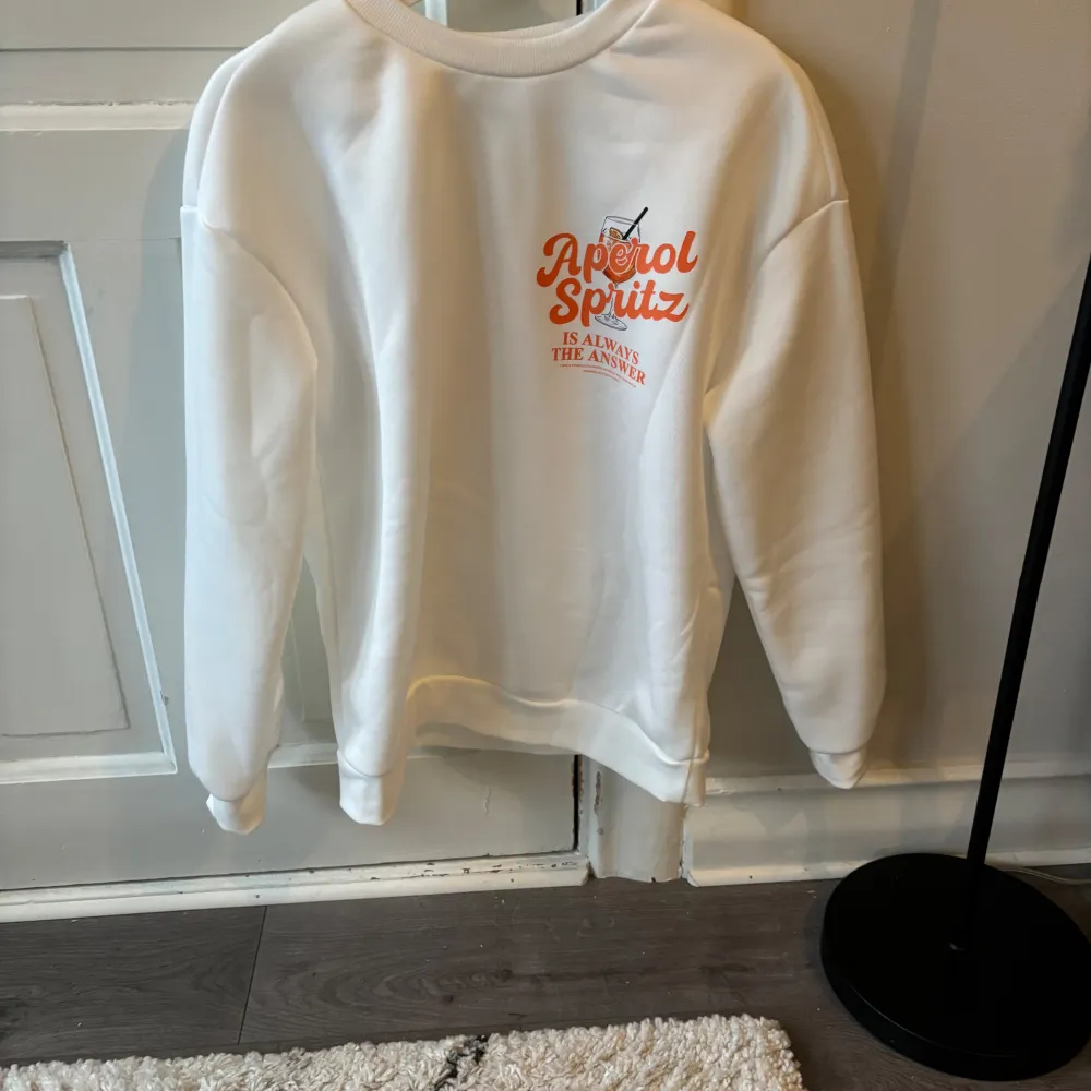 Säljer en vit sweatshirt med ett färgglatt Aperol Spritz-tryck i orange på både fram- och baksidan. Tröjan har långa ärmar och en rund halsringning. Perfekt för en avslappnad stil. Helt ny strl 34/36. Neuletakit & Villapaidat.