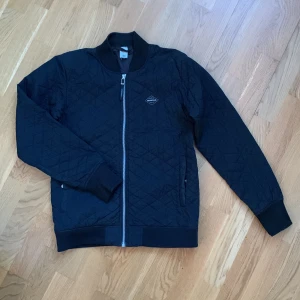 Svart quiltad bomberjacka från Didriksons - Snygg svart quiltad bomberjacka från Didriksons med dragkedja framtill. Jackan har en klassisk bomberkrage och långa ärmar. Perfekt för en stilren look.