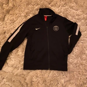Svart träningsjacka PSG - Säljer en svart PSG träningsjacka från Nike. Perfekt för sportiga aktiviteter eller daglig användning. Har endast använt i skolan och ej under träning. 