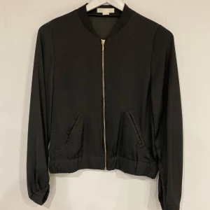 Mörkgrön bomberjacka från H&M - Snygg mörkgrön bomberjacka från H&M med dragkedja framtill. Jackan har långa ärmar och en klassisk bomberstil med muddar vid ärmslut och nederkant. Jackan är väldigt tunn och är därför bäst lembad till dommaren eller bara för att piffa till en outfit när man inte behöver vara så varm