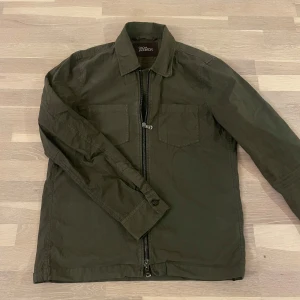 Overshirt från Oscar Jacobson - Snygg olivgrön overshirt från Oscar Jacobson med dragkedja framtill. Jackan har en klassisk krage och två bröstfickor. Perfekt för en stilren look under vår och sommar. NYPRIS 2599 kr.