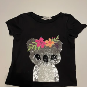 Svart t-shirt med paljettkoala från H&M - Söt svart t-shirt från H&M med en glittrig koala i paljetter på framsidan. Koalan har en färgglad blomsterkrans i rosa, blått och gult. Vändbara paljetter för variation! 