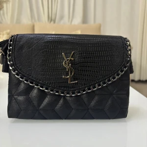 Svart axelväska med kedjedetalj - Snygg svart axelväska från YSL med quiltad design och elegant kedjedetalj. Väskan har en texturerad klaff och ett metalliskt YSL-emblem på framsidan. Perfekt för att ge en lyxig touch till din outfit.