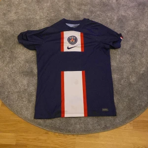 Blå PSG fotbollströja med Sergio Ramos nummer 4 - Snygg blå PSG fotbollströja från Nike med korta ärmar och Sergio Ramos namn och nummer 4 på ryggen. Tröjan har ett vitt och rött vertikalt mönster på framsidan och PSG-loggan på bröstet. Perfekt för fans av Paris Saint-Germain och Sergio Ramos!