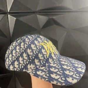 Mönstrad keps från Dior - Snygg keps från Dior med ett blått och vitt mönster. Kepsen har en broderad gul logotyp framtill och är tillverkad i ett bomullsmaterial. Perfekt för att ge din outfit en lyxig touch. Använd runt 5-10 gånger. Den har ett band som blir perfekt att justera storlek på kepsen.
