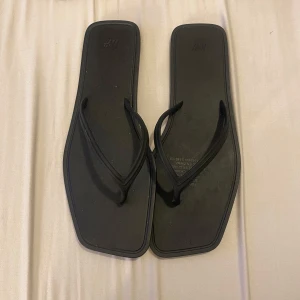 Svarta flip-flops från H&M - Säljer ett par svarta flip-flops från H&M. De har en fyrkantig form och är perfekta för sommardagar. Tillverkade i syntetmaterial med en enkel design.