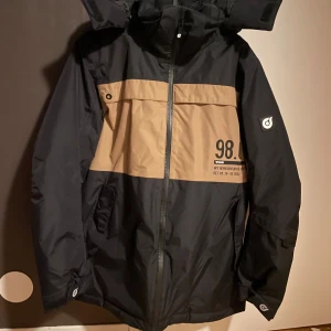 Svart och beige jacka med huva - Snygg svart och beige jacka från Kappahl  med huva och dragkedja. Jackan har en cool design med tryck på bröstet och en praktisk ficka med knapp. Perfekt för kyligare dagar.