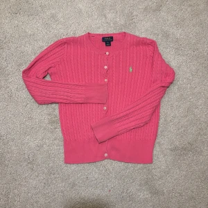 Rosa kofta från Polo Ralph Lauren - Säljer en snygg rosa kofta från Polo!❣️Ralph Lauren med kabelstickat mönster och grönt broderat logotyp på bröstet. Koftan har långa ärmar och knappar framtill💘. Perfekt för en stilren look!💕Säljer då den är för liten på mig som har 158/164 barnstorlek!❣️ 