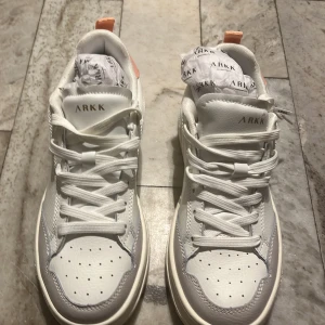 Vita sneakers från ARKK - Snygga vita sneakers från ARKK med detaljer i ljusrosa. Skorna har snörning och en modern design. Perfekta för en stilren look. Ej använda. Liten i storleken 
