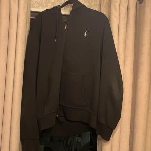 Svart hoodie från Polo Ralph Lauren - Säljer en klassisk svart hoodie från Polo Ralph Lauren med dragkedja och huva. Den har en broderad logotyp på bröstet och praktiska fickor framtill. Perfekt för en avslappnad stil.
