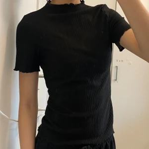 Svart ribbad topp - svart ribbad topp med korta ärmar och en subtil volangdetalj vid halsen. Aldrig använd från märket ONLY. Original pris var 180 kr, säljer för 90 kr 