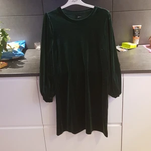 Mörkgrön sammetsklänning från Gina Tricot - Elegant mörkgrön klänning i mjukt sammetstyg från Gina Tricot. Klänningen har långa ärmar och en rund halsringning, perfekt för en stilren look.