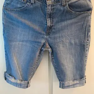 Säljer ett par klassiska jeansshorts från Levi's i en snygg blå färg. De har en normal passform och är tillverkade i slitstarkt denim. Shortsen har fem fickor och en uppvikt kant för en avslappnad look. Perfekta för sommaren!