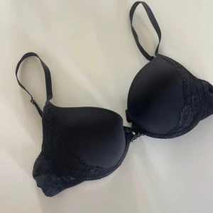 Svart bh med spetsdetaljer - Snygg svart bh med vadderade kupor och spetsdetaljer längs kanterna. Den har justerbara axelband och en liten rosett mellan kuporna. 