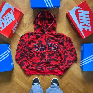 Röd camo hoodie från BAPE - Säljer en supercool röd camo hoodie från BAPE. Den har en fet camouflagemönster i rött och svart med BAPE-loggan broderad på framsidan. Hoodien har dragkedja och är perfekt för höst och vinter. Den är i nyskick och riktigt skön att ha på sig. Perfekt för streetwear-stilen!