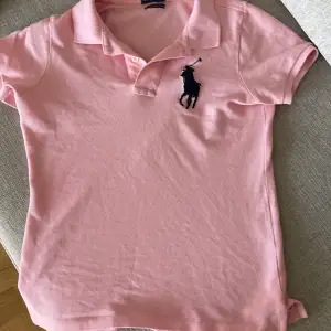 Säljer en snygg rosa pikétröja från Ralph Lauren. Den har en klassisk krage och korta ärmar, perfekt för en avslappnad men stilren look. Tröjan är i modellen 'The Skinny Polo' och har det ikoniska broderade logotypen på bröstet. Passar perfekt till både vardag och lite finare tillfällen!