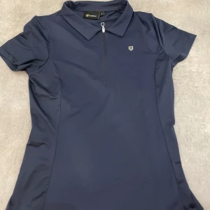 Mörkblå ridtröja Fairfield  - Snygg mörkblå polotopp med korta ärmar och dragkedja framtill. Perfekt för en sportig look. Den har en liten logga på bröstet och är tillverkad i ett bekvämt material.