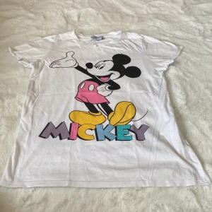 Vit Disney t-shirt med Musse Pigg - Säljer en vit t-shirt från Disney med ett stort tryck av Musse Pigg i färgerna svart, rosa, gul och blå. T-shirten är kortärmad och har en avslappnad passform. Perfekt för alla Disney-fans! 🐭