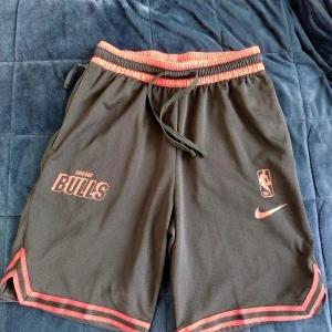 Chicago Bulls shorts från Nike - Snygga svarta Chicago Bulls Courtside Nike NBA-shorts med röd detaljering och Dri-FIT material. Perfekta för basket eller träning. Elastisk midja med snörning för bästa passform.