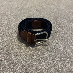 Svart flätat bälte med bruna detaljer - Snyggt svart flätat bälte med bruna skinnliknande detaljer och ett silverfärgat spänne. Perfekt för att ge en stilren touch till din outfit.