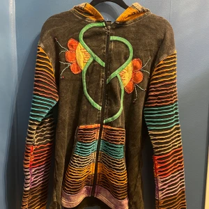 Färggkad hippie jacka med dragkedja - Säljer en unik jacka med dragkedja och huva. Tröjan har färgglada ränder längst armarna, fickorna och luvan i utstickande tyg. Tröjan är väldigt bra kvalitet även fast den är köpt second hand, 100% bomull, säljer då jag inte använder den längre, storlek L. 
