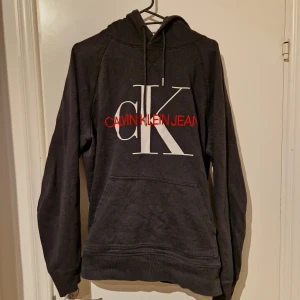 Grå hoodie från Calvin Klein - Säljer en snygg grå hoodie från Calvin Klein med stor vit logga och röd text på framsidan. Tröjan har en klassisk känguruficka och justerbar huva med snören. Perfekt för en avslappnad stil.