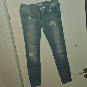 Blå slitna jeans - Säljer ett par blå jeans med slitningar för en trendig look. De har en dragkedja och knappar framtill. Perfekta för en avslappnad stil.