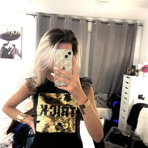 Svart topp med guldpaljetter - Trendig svart topp med glittriga guldpaljetter framtill och transparenta ärmar med prickigt mönster. Perfekt för en kväll ute eller en festlig tillställning. Toppen har en tight passform och ärmlös design.