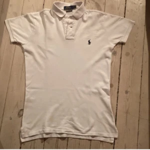 Vit pikétröja från Ralph Lauren - Säljer en klassisk vit pikétröja från Ralph Lauren i custom slim fit. Tröjan har korta ärmar och en broderad logotyp i blått på bröstet. Perfekt för en stilren look. Hör av dig vid intresse!