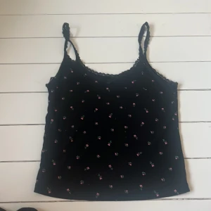 Svart blommig linnetopp från H&M Divided - Säljer en svart linnetopp från H&M Divided med små rosa blommor och spetsdetaljer. Toppen har smala axelband och en söt design som passar perfekt för sommardagar. Perfekt för att ge din outfit en romantisk touch 🌸