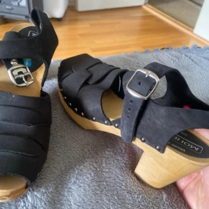  träskor  - Snygga svarta träskor från Moheda med träklack och justerbart spänne. Skorna har en klassisk design med öppen tå och remmar i syntetmaterial. Perfekta för en stilren look.