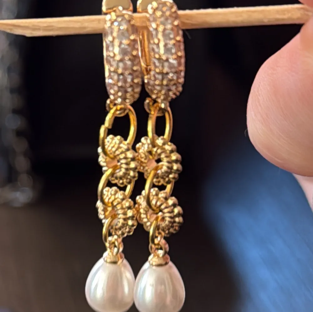 Eleganta guldörhängen med glittriga detaljer och hängande vita pärlor. Perfekta för att ge en lyxig touch till din outfit. De har en vacker design med små guldfärgade blommor och stenar.. Asusteet.
