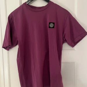 Lila t-shirt från Stone Island - Säljer en snygg lila t-shirt från Stone Island med korta ärmar och en klassisk rund halsringning. T-shirten har en broderad logotyp på bröstet som ger en stilren look. Perfekt för en avslappnad stil.