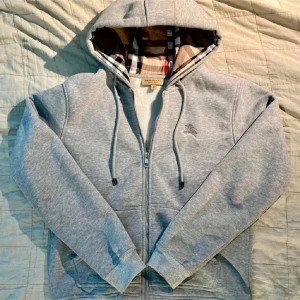  Burberry zip hoodie - Säljer en stilren grå hoodie från Burberry med klassiskt rutigt mönster i huvan. Tröjan har dragkedja framtill och en broderad logga på bröstet. Perfekt för en avslappnad och trendig look. Hör av dig vid intresse!