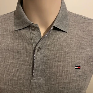 Grå pikétröja från Tommy Hilfiger - Snygg grå pikétröja från Tommy Hilfiger med klassisk krage och knappslå. Tröjan har korta ärmar och det ikoniska Tommy Hilfiger-logotypen broderad på bröstet och ärmen. Perfekt för en stilren och avslappnad look.