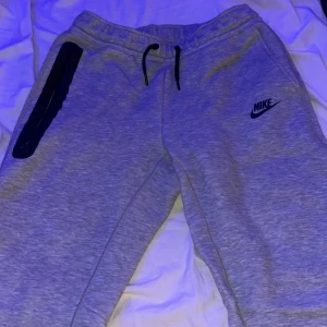 Grå Nike tech byxor  - Snygga grå mjukisbyxor från Nike med svart logga på benet. Byxorna har en praktisk dragkedja på sidan och snörning i midjan för bästa passform. Perfekta för en avslappnad stil. ( Det är storlek 158 men det passar XS )