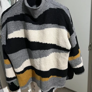 Randig stickad tröja från Vila - Säljer en mysig stickad tröja från Vila med ett randigt mönster i svart, grått, vitt och gult. Tröjan har en hög krage och långa ärmar, perfekt för kyliga dagar. Den är både stilren och bekväm.