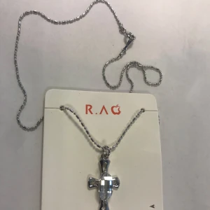 Silverfärgat halsband med kors  - Elegant silverfärgat halsband från R.A.G med ett korshänge. Kedjan är tunn och smidig, perfekt för att bära till olika tillfällen. Korset har en stilren design som ger en tidlös känsla.  Helt ny 