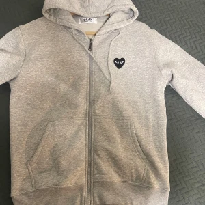 Grå hoodie från Comme des Garçons Play - Säljer en grå hoodie från Comme des Garçons Play med det ikoniska hjärtmärket på bröstet. Tröjan har dragkedja och en stor ficka framtill. Perfekt för en casual look. 