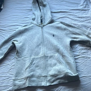 Grå hoodie från Ralph Lauren - Säljer en stilren grå hoodie från Ralph Lauren med dragkedja och klassisk logga på bröstet. Perfekt för en avslappnad look. Den har en praktisk ficka framtill och en bekväm huva.