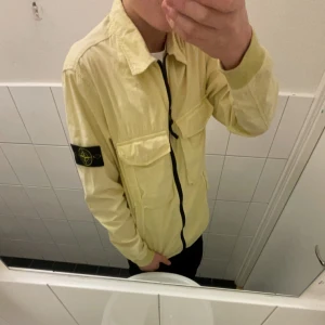 Gul overshirt från Stone Island - Säljer en stilren gul overshirt från Stone Island med dragkedja och två bröstfickor. Den har en ikonisk patch på ärmen och är perfekt för lager-på-lager-stil. Passar bra till både vardag och mer avslappnade tillfällen.