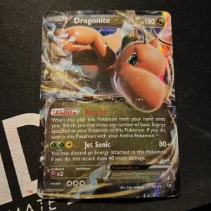 Dragonite Pokémon-kort - Säljer ett Dragonite Pokémon-kort med 180 HP. Kortet har förmågan 'Bust In' och attacken 'Jet Sonic'. Det är ett holografiskt kort med en färgglad design och visar Dragonite i en dynamisk pose. Perfekt för samlare eller Pokémon-entusiaster!