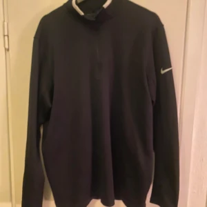 Svart långärmad tröja från Nike - Säljer en stilren svart långärmad tröja från Nike med en diskret logga på ärmen. Perfekt för träning eller vardagsbruk. Tröjan har en dragkedja vid halsen för justerbar komfort.