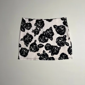 Vit kortkjol med svart blommönster - Trendig vit kortkjol med stora svarta rosor. Perfekt för att ge en edgy touch till din outfit. Kjolen har en rak passform och är tillverkad i ett mjukt material som ger en bekväm känsla.