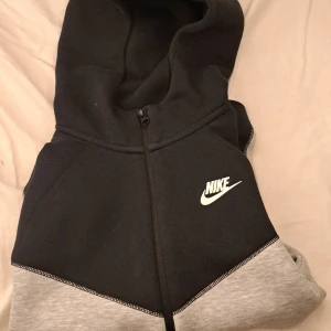 Svart och grå hoodie från Nike - Säljer en snygg svart och grå hoodie från Nike med dragkedja framtill. Tröjan har en klassisk design med huva och två fickor med dragkedjor. Perfekt för en avslappnad stil.