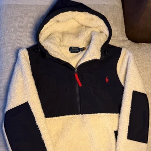 Vit och marinblå fleecejacka från Ralph Lauren - Säljer en snygg vit och marinblå fleecejacka från Ralph Lauren. Jackan har en dragkedja framtill och en huva. Den är dekorerad med Ralph Laurens ikoniska logga i rött på bröstet. Perfekt för kyliga dagar!