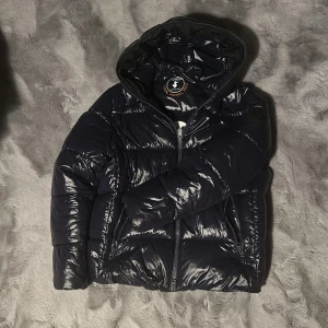 Svart pufferjacka från Save the Duck - Säljer en mörkblå pufferjacka från Save the Duck. Jackan är 100% djurfri och har en glansig finish med dragkedja framtill. Den har en huva och är perfekt för kyligare väder. Jackan är knappt använd och därför i 10/10 skick. 