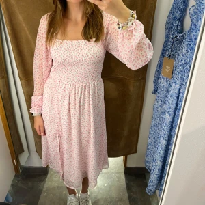 Rosa blommig klänning - Charmig rosa klänning med blommigt mönster och långa ärmar. Den har en smickrande passform med elastisk midja och en luftig kjol som slutar under knäna. Perfekt för en somrig look!