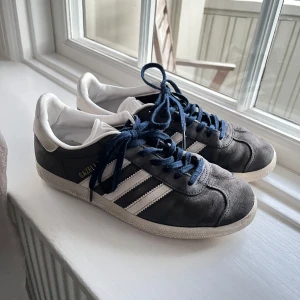 Adidas Gazelle sneakers - Snygga blåa Adidas Gazelle sneakers med vita detaljer och mörkblå snörning. Skorna har den klassiska tre-randiga designen och en bekväm sula. Perfekta för en stilren och sportig look.